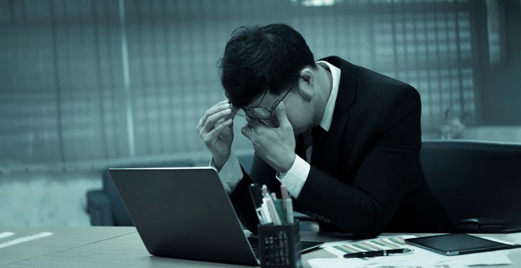 A stressed businessman in a black suit, frustrated with his mistakes, sitting at a desk with his head in his hands.